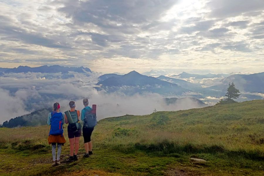 Drei Wandernde auf einer Bergwiese mit Blick auf Wolken und umliegende Berge